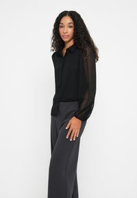 Chemise noire à boutons avec des manches en tulle à pois, associée à un pantalon gris taille haute à jambes larges. Texture lisse, design minimaliste.