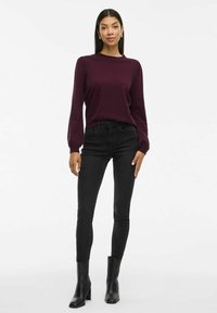 Burgundská dlouhý rukáv top, kombinovaný s černými skinny džínami. Modelka má na sobě černé zúžené kotníčkové boty na podpatku, které zdůrazňují elegantní, přiléhavý siluetu.