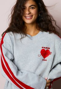 Siva majica z rdečimi črtami na rokavih, z rdečim vezenim napisom "PARIS AMOUR" in rdečim grafičnim srcem. Mehki bombažni material.