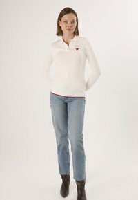 Witte poloshirt met lange mouwen en een rood hartlogo, aansluitend ontwerp, geribbelde kraag en mouwen. Gecombineerd met lichtblauwe rechtgesneden jeans en zwarte laarzen.