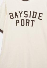 Chemise crème à manches courtes avec un texte brun foncé "BAYSIDE PORT" imprimé sur la poitrine et des bordures assorties brunes sur les manches et le col.