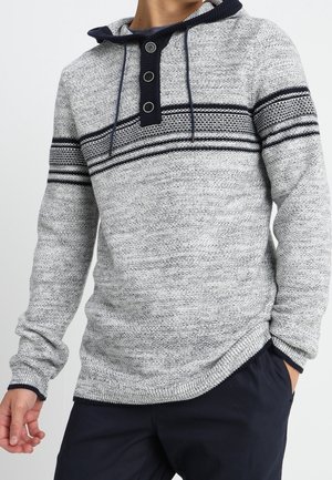 Šedá pletená mikina s kapucí s tmavě modrými vodorovnými pruhy přes hrudník a paže, tři tmavé knoflíky na légu, nošená s tmavými kalhotami.