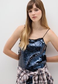 Schwarzes und blaues Pailletten-Trägershirt mit dünnen Trägern, das eine glänzende Textur aufweist. Mit einem gebundenen karierten Flanellhemd in der Taille. Schlichter Hintergrund.