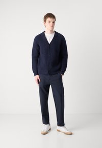 Μπλε πλεκτή ζακέτα με V λαιμόκοψη, κουμπί κλεισίματος, ραβδωτή υφή, συνδυασμένη με σκούρα παντελόνια και λευκά αθλητικά. Απλός σχεδιασμός.