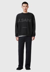 Jeune homme portant un sweat à manches longues noir avec le texte "ALLSAINTS LONDON", un pantalon noir et des chaussures noires, se tenant devant un fond gris.