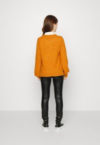 Pullover de malha laranja com um corte solto, punhos canelados, usado sobre uma camisa branca de colarinho, combinado com calças de couro pretas e ténis brancos.