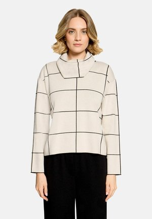 Femme aux cheveux blonds courts porte un haut blanc à manches longues avec des lignes noires en grille et un large col entonnoir, associé à un pantalon noir.