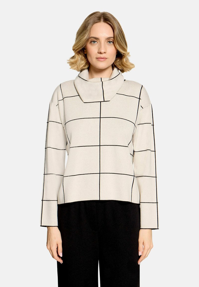 Femme aux cheveux blonds courts porte un haut blanc à manches longues avec des lignes noires en grille et un large col entonnoir, associé à un pantalon noir.
