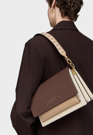 Persona con abrigo marrón oscuro que lleva un bolso de hombro TOUS tricolor con correa beige y detalle de cadena dorada, visto desde atrás.