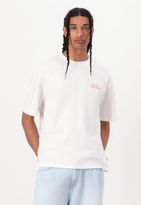 Hvit bomulls t-skjorte med korte ermer, med oransjefarget brodert tekst på venstre brystområde, paret med lyseblå denimshorts.