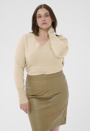 Jeune femme aux cheveux bruns portant un pull beige à encolure en V et une jupe en cuir olive, posant avec une main touchant son cou.