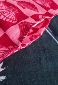 Rot- und rosa kariertes Stoffmuster mit Muscheln; der gefaltete Rand zeigt einen schwarzen strukturierten Hintergrund und erzeugt so ein geschichtetes Erscheinungsbild.