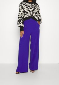 Pull en noir et blanc à motif géométrique avec des manches bouffantes, associé à un pantalon violet ample taille haute avec des poches latérales.