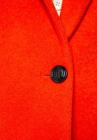 Cappotto di lana arancione brillante con un unico grande bottone nero chiuso vicino al centro.