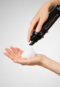 Flacon pulvérisateur noir distribuant une mousse blanche dans une main. Le flacon est muni d'un embout effilé et d'une étiquette avec texte en blanc et rouge. Texture de mousse lisse.