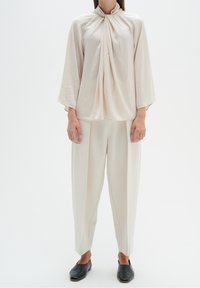 Blusa drappeggiata in tonalità neutre con colletto alto, abbinata a pantaloni larghi leggermente plissettati e scarpe nere slip-on.