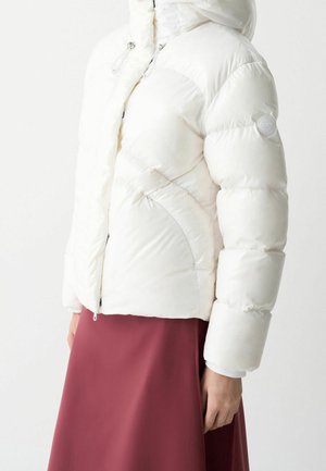 Doudoune blanche à fermeture éclair frontale et capuche, portée avec une jupe rouge foncé, vue de profil des épaules jusqu'aux hanches.