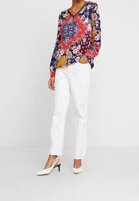 Kuvioitu ja värikäs pusero pitkillä hihoilla, V-pääntie ja kukkakuviot punaisena, sinisenä ja keltaisena. Yhdistetty valkoisiin suoriin housuihin ja korkokenkiin.