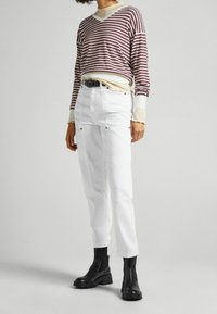 Chemise à manches longues rayée en bordeaux et crème, portée sur un haut crème, associée à un pantalon blanc avec des détails à boutons-pression et des bottines noires.