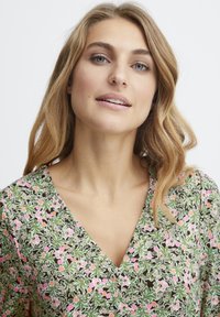 Blumenmusterkleid mit tiefem V-Ausschnitt, das grüne Blätter und rosa Blumen auf einem dunklen Hintergrund zeigt. Weicher Stoff, lockere Passform und kurze Ärmel.