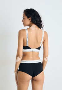 Kvinne med vitiligo merker på huden, iført svart Calvin Klein bralette og undertøy, vender bort mot en enkel lys bakgrunn.