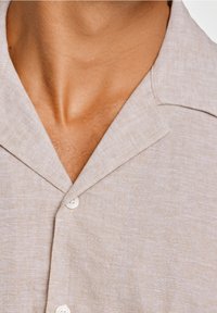 Chemise en lin beige clair avec col, dotée d'une fermeture à boutons et d'un motif texturé subtil. Accent sur la zone du cou et les détails du tissu.