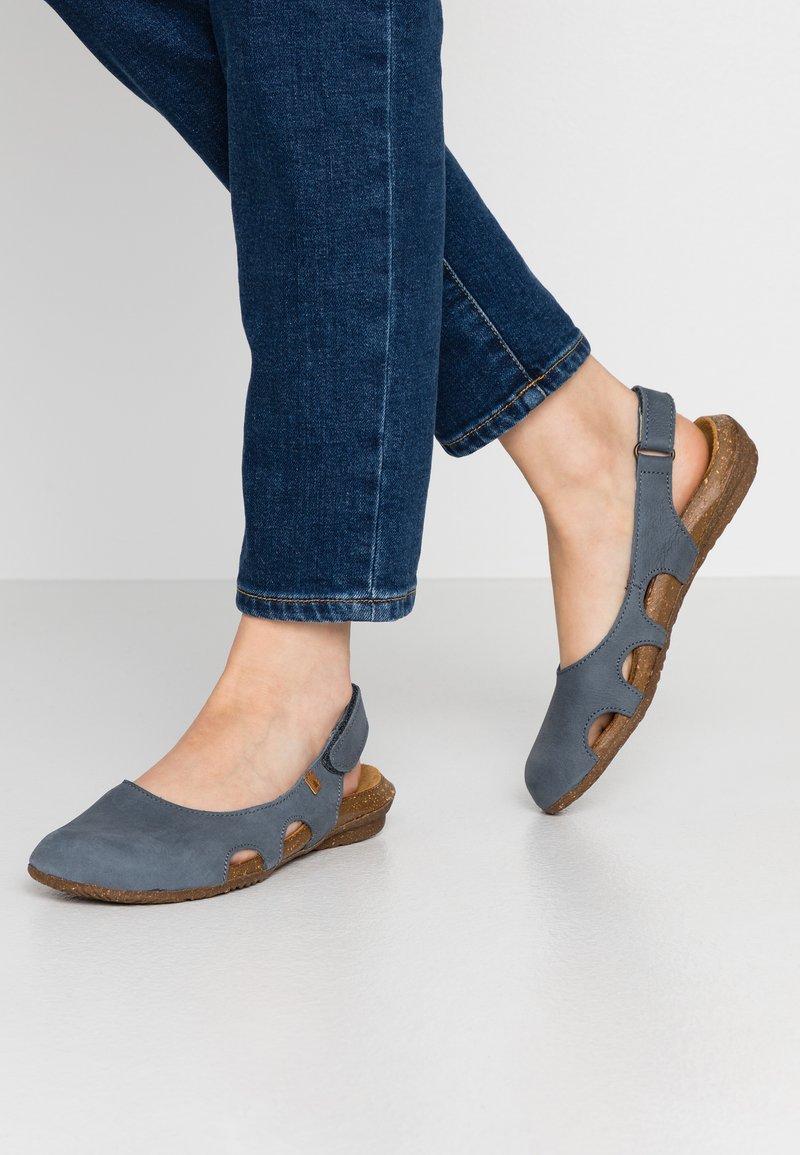 Sandales à talon slingback en cuir gris avec des découpes, une semelle texturée marron et un talon compensé bas, associées à un jean bleu foncé.