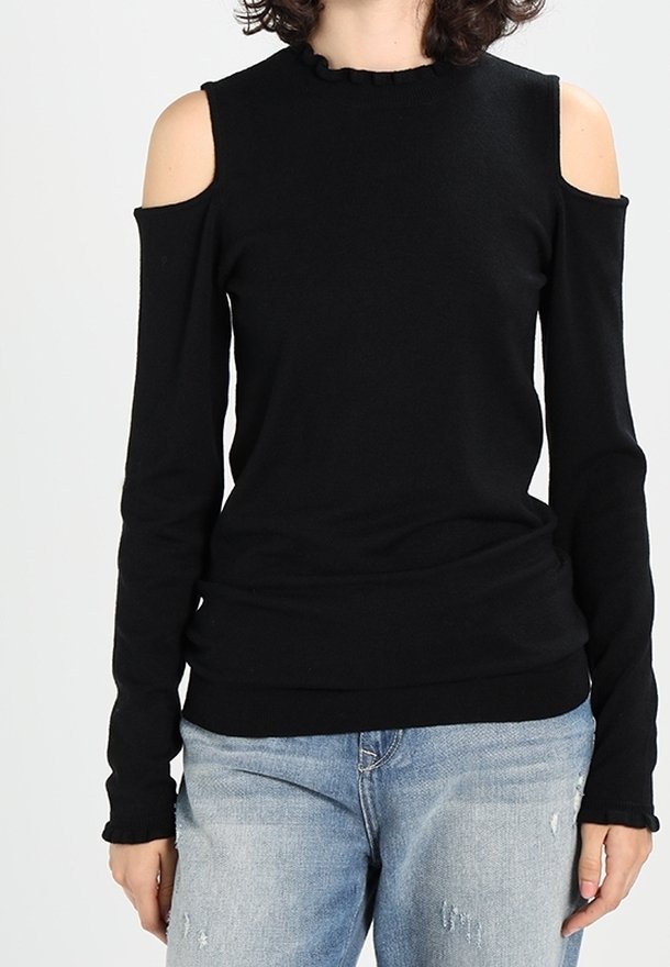 Femme portant un haut noir à manches longues avec épaules dénudées et un jean bleu clair déchiré, debout devant un fond uni.