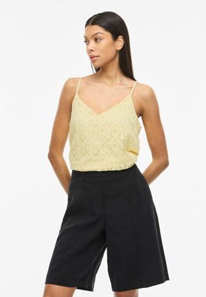 Žena s dugom tamnom kosom nosi žuti čipkani top bez rukava s tankim naramenicama i crne kratke hlače visokog struka do koljena.