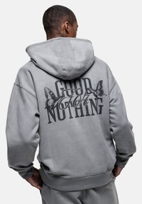 Felpa grigia con cappuccio realizzata in tessuto morbido, dotata di testo nero "GOOD FOR NOTHING" e grafiche di farfalle sul retro. Vestibilità ampia.