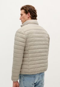 Beige puffer jacket with quilted texture, high collar, and elastic cuffs. Paired with light blue denim jeans. Rear view of model.