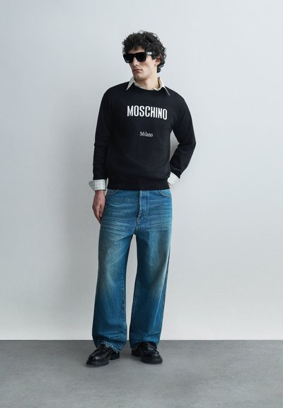 Homme portant un sweat-shirt noir Moschino, une chemise claire, un jean large bleu, des chaussures noires et de grandes lunettes de soleil noires, debout contre un mur gris.