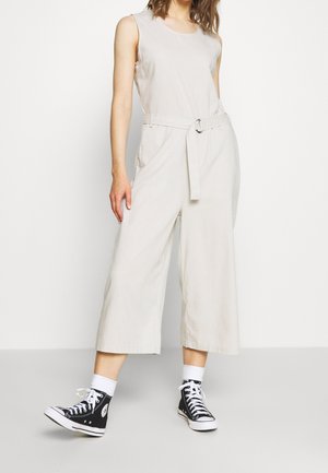 Kobieta ubrana w beżowy kombinezon bez rękawów z paskiem w talii oraz czarne wysokie trampki z białymi skarpetkami.