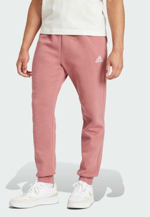 Pantalones de chándal rosas con puños elásticos, bolsillos laterales y un pequeño logo blanco de Adidas en el muslo izquierdo. Combinados con zapatillas blancas.