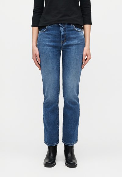 Personne portant un jean bleu taille mi-haute à jambes droites, un haut noir à manches longues et des bottines noires, debout devant un fond uni.