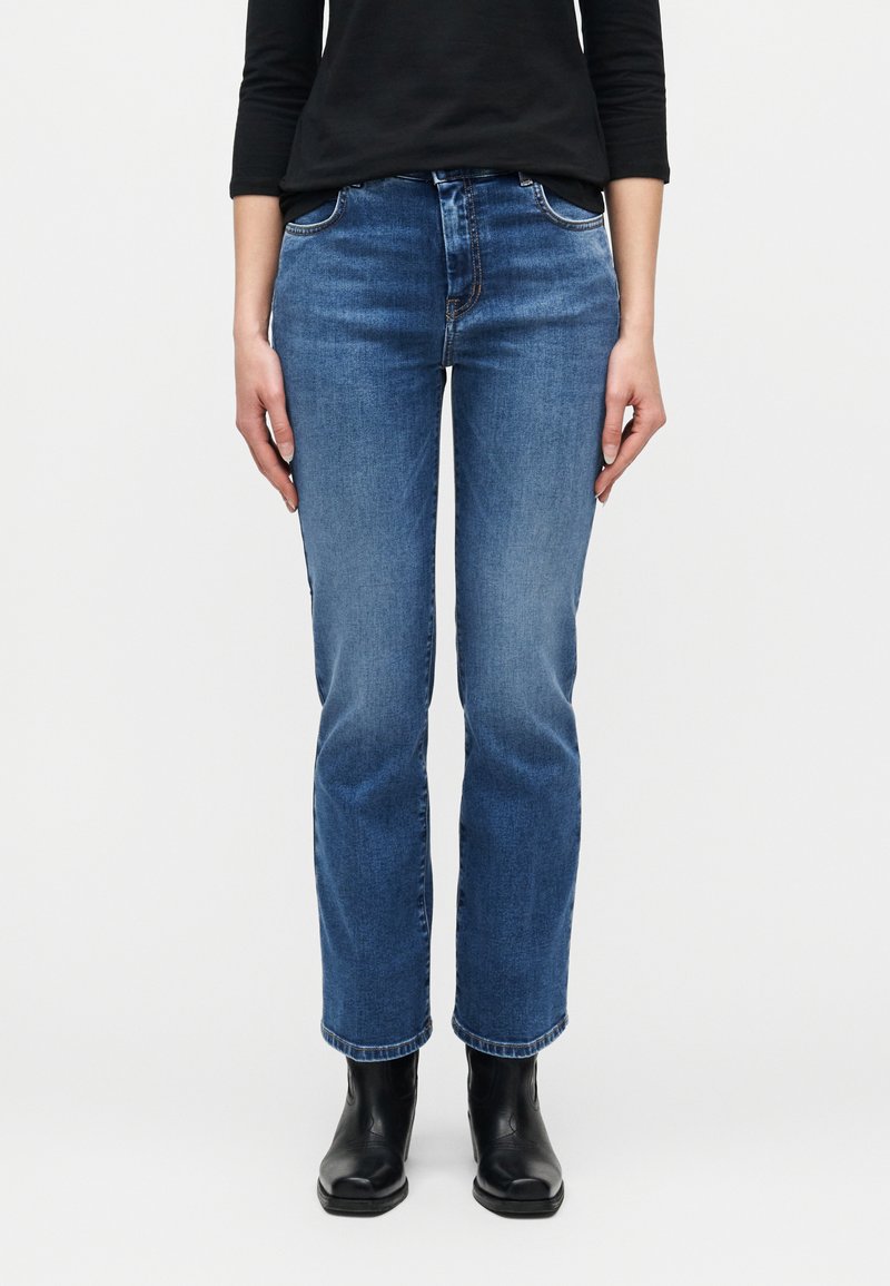 Personne portant un jean bleu taille mi-haute à jambes droites, un haut noir à manches longues et des bottines noires, debout devant un fond uni.