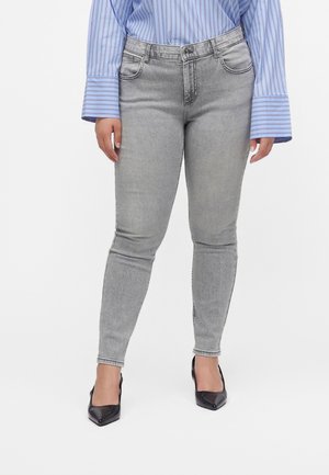 Person iført lysegrå tætsiddende jeans, blå og hvide stribede langærmede skjorte og sorte, spidse hæle, der står mod en hvid baggrund.
