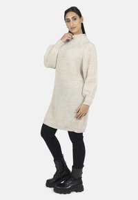 Stickad krämfärgad klänning med hög krage och ribbad textur, kombinerad med svarta leggings och kraftiga svarta snörkängor.