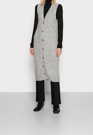 Frau trägt eine ärmellose, knielange graue Strickjacke mit Knöpfen über einem schwarzen Rollkragenpullover, schwarze Hose und schwarze Stiefeletten.