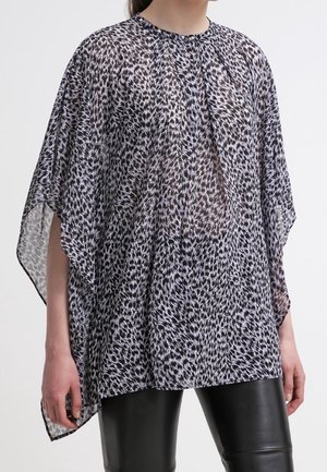 Femme portant un chemisier ample, transparent à imprimé animal noir et blanc avec de larges manches, sur un pantalon en cuir noir.