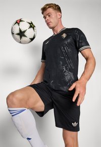 Maillot de football noir à motif floral, rayures blanches sur les manches, associé à un short noir et des protège-tibias blancs ; ballon de football en suspension au-dessus.