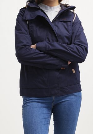 Personne portant une veste à capuche bleu marine par-dessus un pull gris à côtes, les bras croisés, associée à un jean bleu.