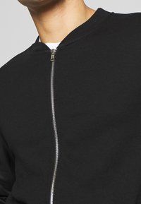 Sudadera negra con cremallera, cuello redondo, textura suave y detalle de cremallera metálica en la parte delantera. Diseño simple y minimalista.