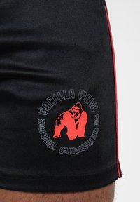 Shorts de sport noirs avec des passepoils rouges sur les côtés. Comprend un logo circulaire rouge représentant un gorille et le texte "GORILLA WEAR" et "Depuis 1982."