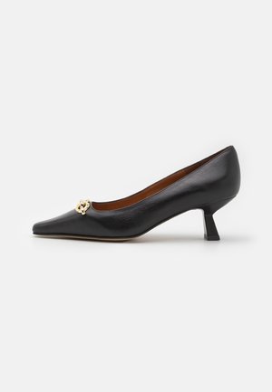 Zwarte leren schoen met spitse neus en lage hak, met een goudkleurig decoratief knoopdetail aan de voorkant, weergegeven op een effen witte achtergrond.