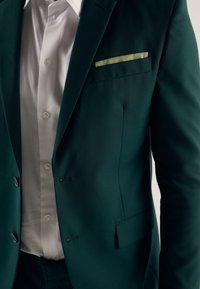 Dunkelgrüne Anzugjacke mit schlanker Passform, einreihigem Knopfverschluss und einem hellgrünen Taschentuch; getragen über einem weißen Hemd.