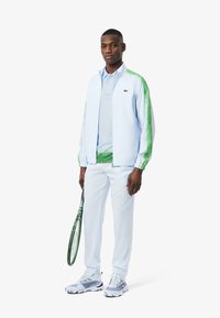 Ljusblå jacka med gröna accenter, kombinerad med en ljusblå polo och byxor. Vita sportskor med ett texturerat mönster, hållande ett tennisracket.