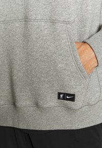 Main dans la poche d'un sweat-shirt gris arborant les logos du Liverpool FC et de Nike sur le bord de la poche avant.