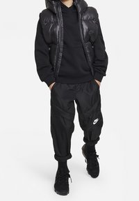 Chaleco de plumas negro sobre una sudadera negra con capucha, combinado con pantalones de chándal negros que presentan paneles texturizados. Zapatillas negras con un acento de logo reflectante.