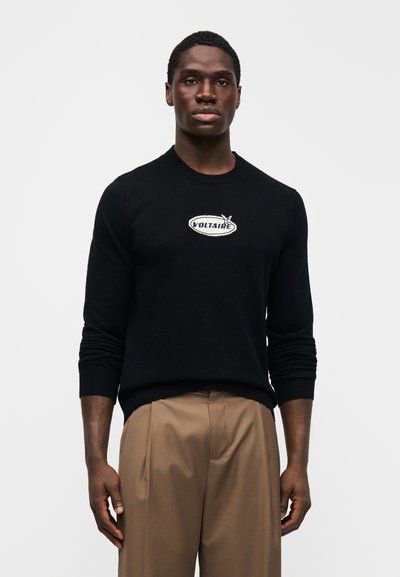 Black knitted sweater with a crew neckline, featuring a white embroidered "VOLTAIRE" logo on a sleek, smooth fabric. Paired with beige trousers.